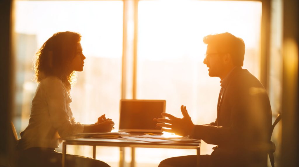 Leidinggevende en medewerker voeren een gesprek aan een tafel met laptop, tegenlicht door raam op de achtergrond