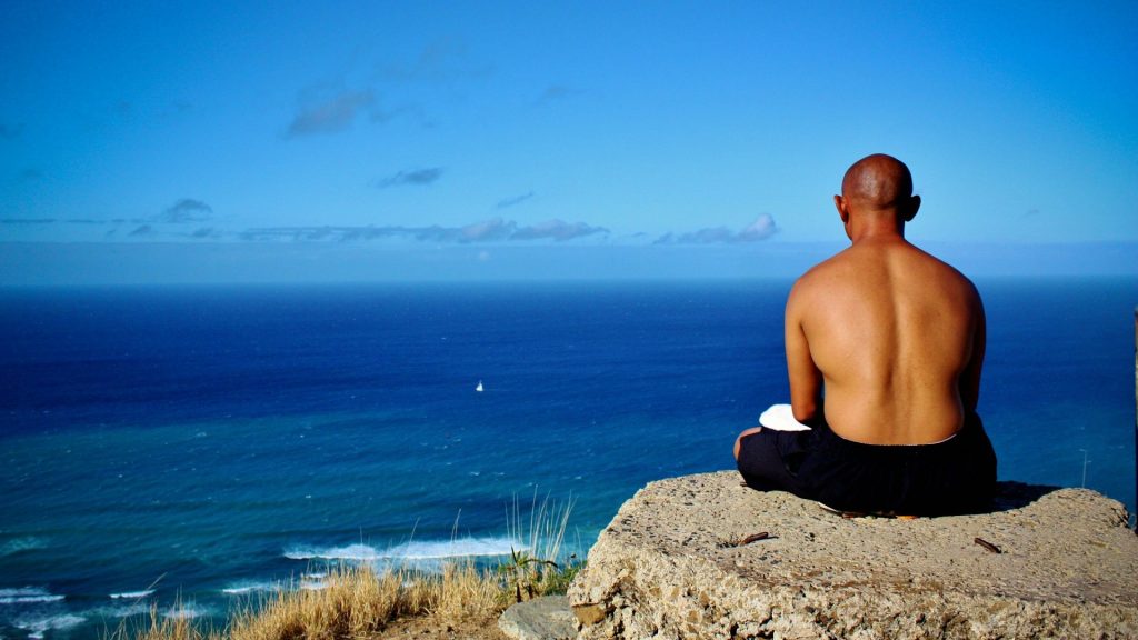 Man mediteert aan zee voor mentale rust, met uitzicht over de kalme oceaan en blauwe lucht