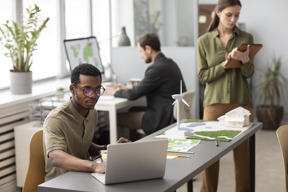 Drie medewerkers in duurzaam kantoor met laptop, windmolenmodel en maquette van een woning.
