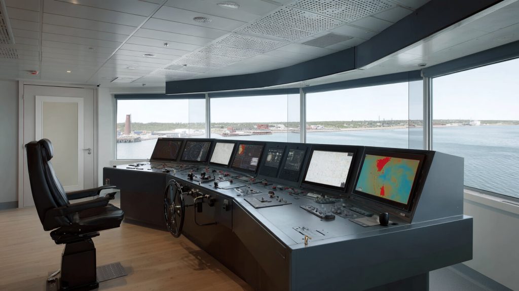 Lege scheepsbrug met stuurstoel en bedieningspaneel vol navigatieschermen, met door grote ramen uitzicht op het water