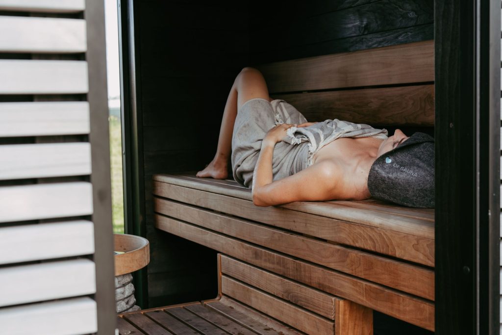Vrouw ligt te ontspannen op een houten bank in een sauna met een saunahoed op.